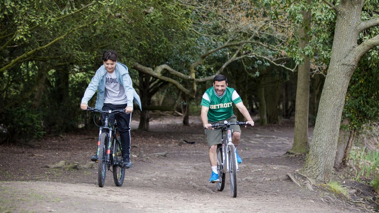 A father and teenage son cycle through woodland with wide pathways on the Osterley Park estate.